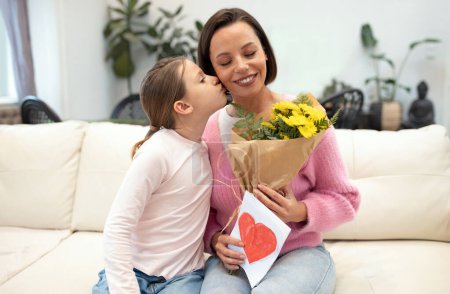 Photo for Happy caucasian small daughter kisses on cheek, congratulates young mother on women day, birthday, holiday gives card and bouquet of flowers in living room interior. Celebrating together at home - Royalty Free Image