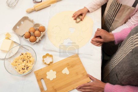 Téléchargez les photos : Les mains de la jeune mère caucasienne et la petite fille dans les tabliers font le biscuit de la pâte avec les formes sur la table de cuisine avec le conseil en bois. Annonce et offre, nourriture maison, confiserie à la maison - en image libre de droit