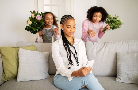 Téléchargez les photos : La fête des mères. Afro-Américaine maman textos au téléphone tandis que ses enfants filles la félicitent en vacances, debout derrière le dos avec des fleurs à la maison. Focus sélectif sur Lady - en image libre de droit