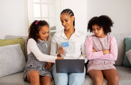 Photo for Displeased Black Preteen Girl Envying Shopping Online With Mother And Sister While Mom Using Laptop And Credit Card Sitting At Home. Family Budget Allocation, Siblings Rivalry And Envy Issue - Royalty Free Image