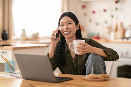 Téléchargez les photos : Joyeux heureux belle jeune femme coréenne dans la cuisine décontractée assis avec ordinateur portable, parler au téléphone, boire du café, manger des cookies, regarder l'espace de copie et sourire, profiter week-end à la maison - en image libre de droit
