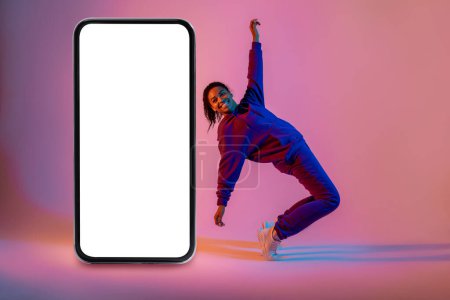 Téléchargez les photos : Danseuse afro-américaine active hip-hop dansant près d'un énorme smartphone avec écran vide sur fond néon rose, espace libre, maquette. Application de danse ou site web - en image libre de droit
