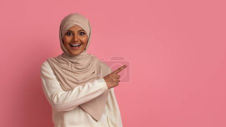 Téléchargez les photos : Regarde ça. Femme musulmane excitée dans le hijab pointant vers l'espace de copie, heureuse femme islamique portant un foulard démontrant la place libre pour la publicité, debout sur fond rose, panorama - en image libre de droit
