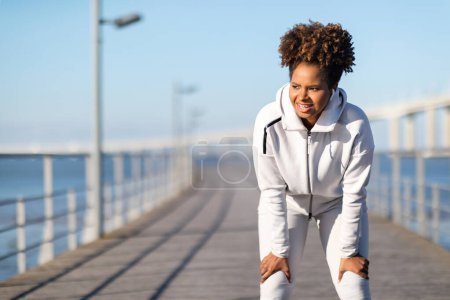 Téléchargez les photos : Fatiguée femme noire sportive prenant souffle après l'entraînement actif en plein air, afro-américaine Jogger en vêtements de sport debout sur la jetée près de la mer, se reposant après la course du matin à l'extérieur, Copier l'espace - en image libre de droit