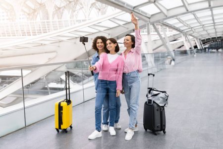 Foto de Tres alegres amigas tomando fotos con selfie stick en el aeropuerto, grupo de mujeres jóvenes felices que viajan juntas, posando en la cámara del teléfono inteligente mientras están de pie con el equipaje en Terminal Hall - Imagen libre de derechos