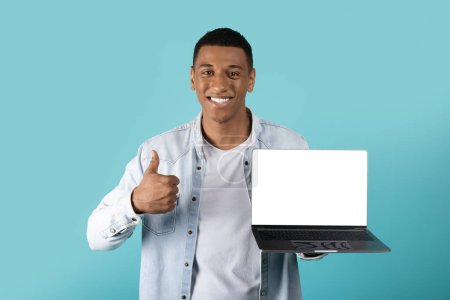 Photo for Cheerful black millennial male showing laptop with blank screen and thumb up isolated on blue studio background. Recommendation app, device, website, presentation and great news - Royalty Free Image