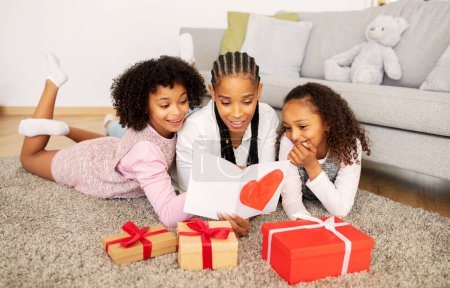 Téléchargez les photos : Mère afro-américaine souriante lisant la carte de vœux recevant des cadeaux de filles précoces pendant les vacances de la fête des mères allongées sur le sol à la maison. Famille Célébration Mommys Anniversaire Concept - en image libre de droit