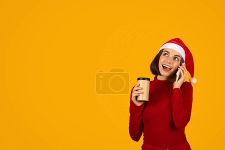 Téléchargez les photos : Jolie jeune femme brune excitée en tenue rouge et chapeau de Père Noël boire du café à emporter, avoir une conversation téléphonique, regarder l'espace de copie avec curiosité et sourire, fond de studio orange - en image libre de droit