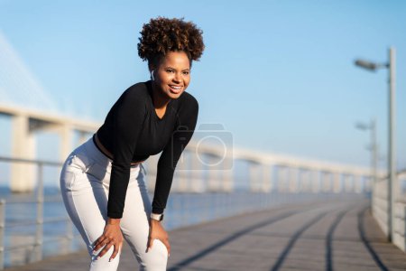 Foto de Sonriente hembra deportiva negra respirando mientras trota al aire libre, hermosa mujer afroamericana en ropa deportiva y auriculares parados en el muelle cerca del mar, descansando después de correr afuera, espacio copiado - Imagen libre de derechos