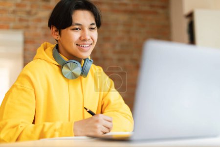 Photo for Remote Education. Happy Korean Guy Using Laptop Computer Learning And Taking Notes Sitting At Desk Indoor. Boy Doing Homework Online At Home. E-Learning Offer Concept. Selective Focus - Royalty Free Image