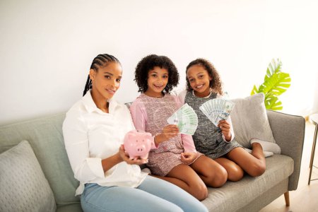 Téléchargez les photos : L'argent familial. Mère afro-américaine et deux filles d'enfant montrant des notes de tirelire et de papier à la caméra élevant des économies ensemble assis sur le canapé à l'intérieur. Grand concept d'offre financière - en image libre de droit