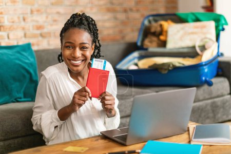 Téléchargez les photos : Femme noire excitée planifiant des vacances, assise avec passeport et billets devant un ordinateur portable et souriant à la caméra, prête pour le voyage, valise déballée sur le canapé - en image libre de droit