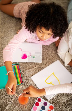 Photo for Vertical Shot Of African American Preteen Girl Drawing With Watercolor In Sketchbook Spending Time With Her Family Lying On Floor At Home On Weekend. Hobby And Leisure. Above View - Royalty Free Image