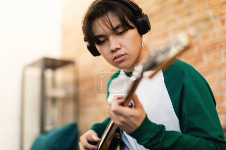Foto de Talentoso adolescente coreano tocando la guitarra eléctrica aprendiendo nuevos acordes con auriculares inalámbricos sentados en el sofá en casa. Guy Aprendiendo a Tocar Canción Favorita en Instrumento Musical. Enfoque selectivo - Imagen libre de derechos