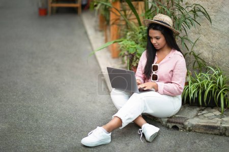 Photo for Smiling serious young black female in casual typing on laptop, looking at hotel, enjoy trip in new city, outside. Device for vacation and journey at summer, travel blog, work and chatting - Royalty Free Image