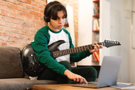 Foto de Asiático adolescente niño jugando guitarra eléctrica con auriculares y el uso de ordenador portátil aprendizaje canción en línea en casa en fin de semana. Music, Technology And Gadgets Concept (en inglés). Enfoque selectivo - Imagen libre de derechos