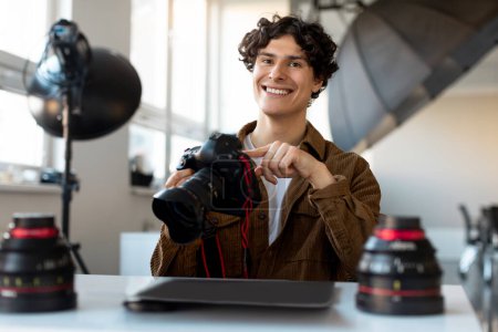 Téléchargez les photos : Photographe masculin joyeux assis sur le lieu de travail dans un photostudio moderne, tenant un appareil photo professionnel et souriant. Emploi et profession concept - en image libre de droit