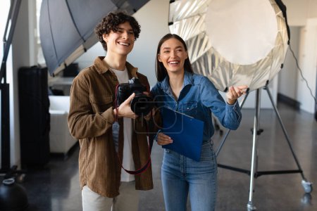 Photo for Young male photographer talking with female art director, talking and preparing for photo shooting, woman pointing forwards and smiling, standing near modern lighting equipment - Royalty Free Image
