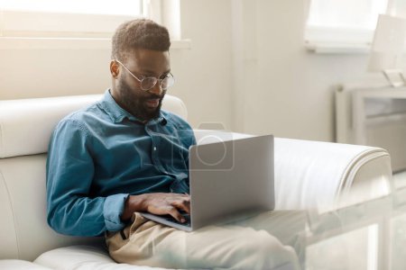 Téléchargez les photos : Jeune homme d'affaires noir assis sur un canapé à l'intérieur du bureau et travaillant sur un ordinateur portable, bavardant avec un client ou participant à une vidéoconférence, copiant de l'espace - en image libre de droit