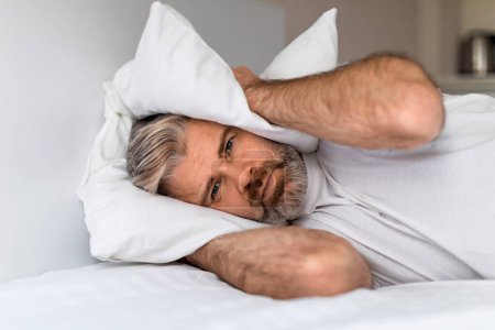 Photo for Portrait of unhappy exhausted middle aged man lying in bed and covering ears with pillow, hearing and suffering from too loud sound or snoring, tired of noisy neighbors. copy space - Royalty Free Image