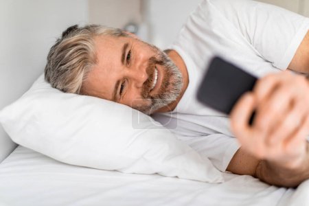 Téléchargez les photos : Délicieux sourire joyeux beau barbu aux cheveux gris homme d'âge moyen en t-shirt blanc couché dans le lit avec téléphone portable dans ses mains, bavarder avec l'amant après le réveil, gros plan - en image libre de droit