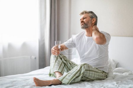 Photo for Unhappy mature man in pain sitting on bed at home, rubbing his neck, suffer from morning muscle strain after sleeping on uncomfortable bed, holding glass of water, looking at copy space - Royalty Free Image
