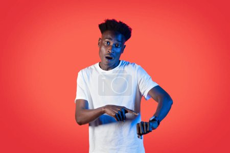 Photo for Timing concept. Overwhelmed shocked young black man in white t-shirt pointing at modern smartwatch on his hand wrist, getting late, red studio background in neon light, copy space - Royalty Free Image