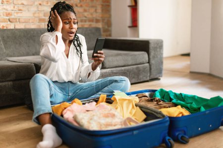 Photo for Traveling problems concept. Shocked black woman using cellphone and reading shocking travel news, sitting near unpacked suitcase at home. Oh no, flight is cancelled - Royalty Free Image