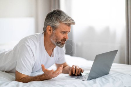 Téléchargez les photos : Colère irritée aux cheveux gris d'âge moyen homme barbu en utilisant un ordinateur portable tout en se relaxant sur le lit à la maison, en regardant l'écran d'ordinateur moderne, en faisant des gestes, en lisant le spam ou avoir des problèmes avec le logiciel, copier l'espace - en image libre de droit