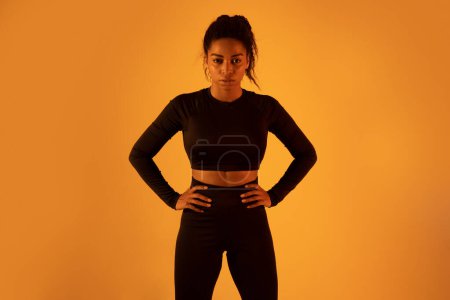 Téléchargez les photos : Portrait de femme de fitness noire portant des vêtements de sport, posant sur fond de studio au néon orange, regardant la caméra. Dame jouissant de son corps mince parfait - en image libre de droit