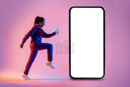Téléchargez les photos : Jeune femme afro-américaine faisant de l'exercice, courant avec un téléphone portable à côté d'un smartphone géant avec écran vide sur fond néon rose, maquette - en image libre de droit