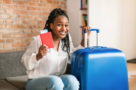 Photo for Cheery african american woman sitting next to suitcase, holding international passport and plane tickets, smiling at camera, getting ready for trip. Summer travel concept - Royalty Free Image