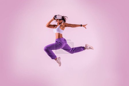 Téléchargez les photos : Jeune femme noire travaillant avec la technologie 3D, sautant en l'air tout en portant un casque de réalité virtuelle, fond studio néon rose. Dame sportive explorant le jeu de fitness virtuel. - en image libre de droit