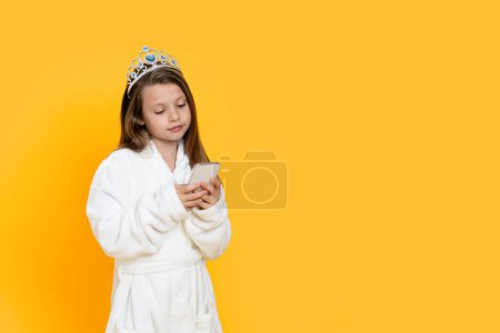 Téléchargez les photos : Petite fille mignonne portant un peignoir et une couronne de jouets à l'aide d'un téléphone intelligent, Preteen Female Child naviguant sur Internet ou faisant des achats en ligne sur un téléphone mobile, debout isolé sur fond jaune, espace de copie - en image libre de droit