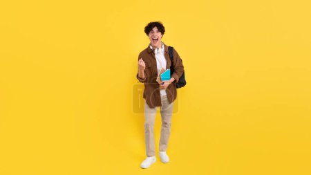 Photo for Emotional Student Guy Shouting Gesturing Yes Celebrating Educational Luck Entering University Posing With Backpack And Notebooks Over Yellow Studio Background, Looking At Camera. Panorama - Royalty Free Image