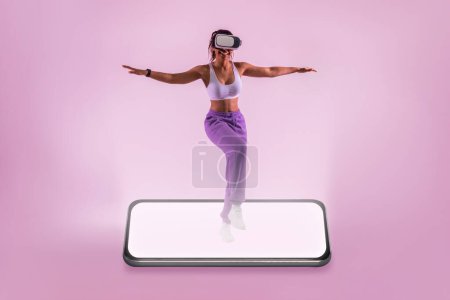 Téléchargez les photos : Application de fitness moderne. Adaptez la femme noire dansant sur l'écran géant de smartphone, s'exerçant pendant l'entraînement en ligne sur le fond rose de studio de néon, espace libre - en image libre de droit