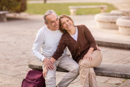 Téléchargez les photos : Joyeux mari aîné et femme étreignant avoir un rendez-vous romantique dans le parc assis sur le banc à l'extérieur. Conjoints retraités profitant de la vie en plein air le week-end. Mariage heureux et retraite - en image libre de droit