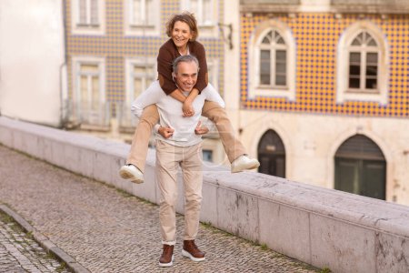 Téléchargez les photos : Positif mature mari portant femme piggyback avoir amusant marcher à l'extérieur. Couple de touristes profitant de vacances à pied à Lisbonne. Happy Senior Life Concept. Prise de vue complète - en image libre de droit