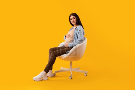 Photo for Motherhood concept. Excited pregnant lady tenderly embracing belly while sitting on chair over yellow studio background. Expectant woman enjoying pregnancy time - Royalty Free Image