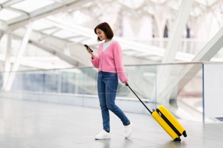 Foto de Registro en línea. Mujer joven sonriente caminando con el equipaje en el aeropuerto y el uso de Smartphone, Mujer feliz Millennial navegando por Internet en el teléfono móvil mientras va a la puerta de vuelo, Copiar el espacio - Imagen libre de derechos