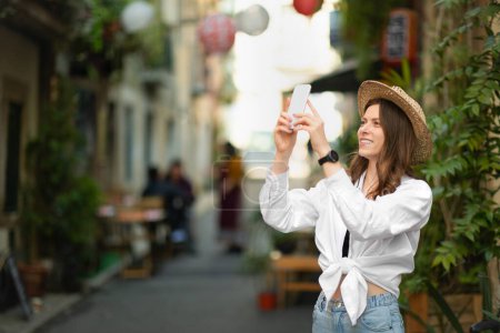 Photo for Inspired happy young caucasian lady in casual and hat makes photo on smartphone looks at sights in new city, outside. Application, blog for vacation and trip in summer, travel blog - Royalty Free Image