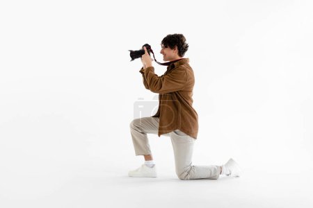 Téléchargez les photos : Jeune photographe masculin en tenue classique, tenant son appareil photo professionnel et prenant des photos, debout sur un genou sur fond de studio blanc, vue latérale - en image libre de droit