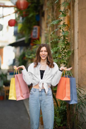 Photo for Laughing young caucasian lady in casual with many bags of purchases enjoy shopping tour in new city, outside, vertical. Fashion vacation and travel in summer, buy clothes and emotions - Royalty Free Image