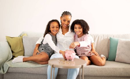 Photo for Great Bank Offer. Happy Black Mother And Her Daughters Putting Coin In Piggybank Raising Money, Hugging Sitting On Couch At Home. Family Savings And Financial Safety Concept - Royalty Free Image