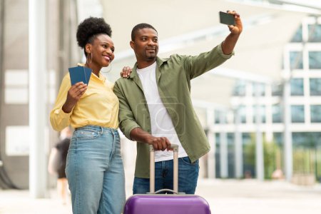 Téléchargez les photos : Vacances Selfie. Joyeux couple de touristes noirs prenant des photos via téléphone portable posant avec valise et passeports debout à l'aéroport en plein air. Concept de transport et tourisme - en image libre de droit