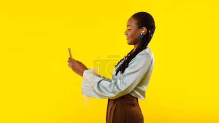Foto de Smiling Black Lady usando Smartphone usando auriculares inalámbricos y escuchando música en línea en el estudio sobre fondo amarillo. Podcast o anuncio de aplicación musical. Panorama, Vista lateral - Imagen libre de derechos