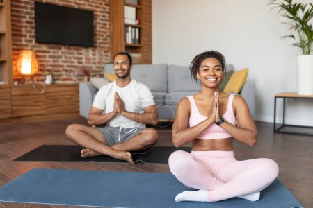 Foto de Feliz joven pareja negra en ropa deportiva haciendo ejercicios, practicando yoga en la estera, disfrutar de la comodidad, la calma y el silencio, el descanso y relajarse en el interior de la sala de estar, espacio de copia. Meditación en casa, cuidado corporal - Imagen libre de derechos