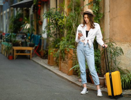 Photo for Serious young caucasian lady in casual and hat with suitcase looks at smartphone map search for attractions in new city, outside. App and blog for vacation and travel in summer, hotel problems - Royalty Free Image