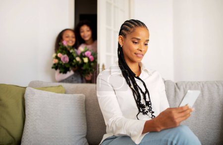 Téléchargez les photos : Mère noire tressée utilisant un téléphone intelligent pendant que ses enfants préparent un cadeau surprise pour elle debout avec un bouquet de fleurs dans la porte le jour de son anniversaire à la maison. Vacances en famille. Focus sélectif - en image libre de droit