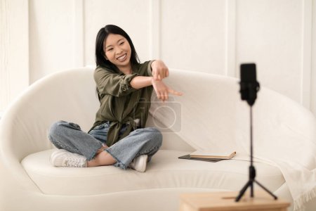 Téléchargez les photos : Célèbre blogueur jeune femme coréenne attrayante streaming de la maison, influenceur féminin assis sur le canapé dans le salon, en regardant la caméra du smartphone et des gestes. Concept de blogging - en image libre de droit